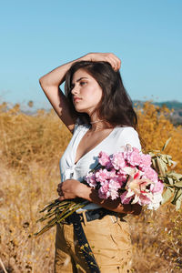 Woman holding flowers while standing on land against clear blue sky