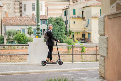 Side view full length young businessman in a suit riding an electric scooter while commuting to work 