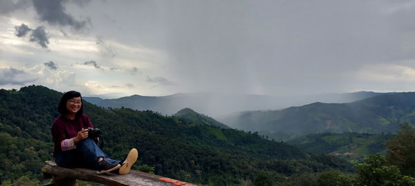 Portrait of smiling young woman sitting on mountain against sky