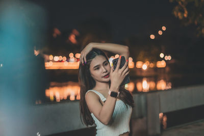 Smiling young woman taking selfie on bridge over river at night