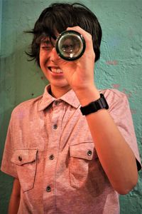 Smiling boy looking through glass bottle while standing against wall