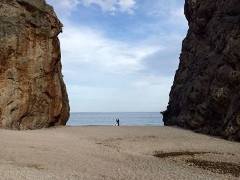 Distant view of wedding couple standing at beach amidst cliffs against sky