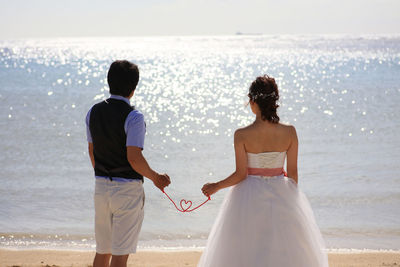 Rear view of couple holding heart shape made with rope while standing at beach