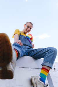 Happy non-binary person with rainbow flag sitting on wall