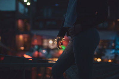 Midsection of woman holding illuminated string light in city at night