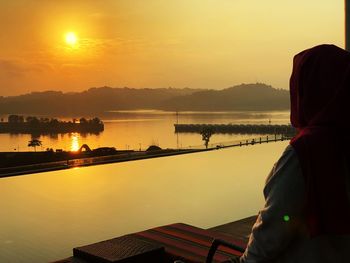 Rear view of man looking at lake against sky during sunset