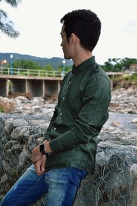 Young man sitting on retaining wall against sky