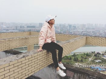 Full length of man sitting on brick wall against city