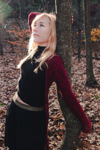 Portrait of young woman standing by tree trunk in forest during autumn