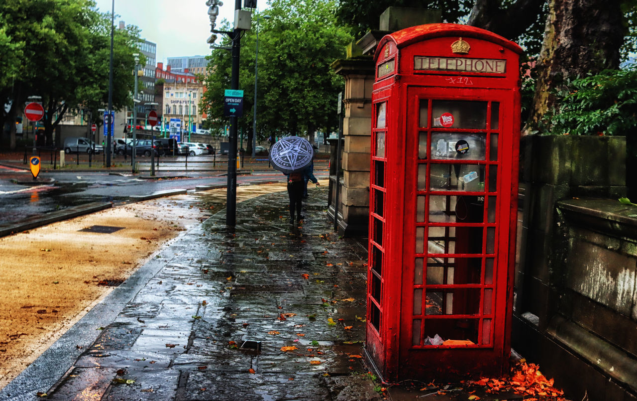 telephone booth, telephone, urban area, communication, city, architecture, street, tree, red, plant, nature, technology, built structure, building exterior, day, payphone, no people, outdoors, footpath, text, sidewalk, road, western script, transportation, public space