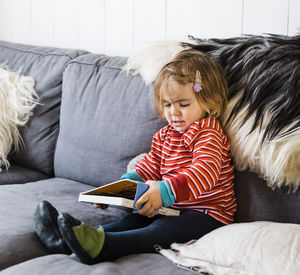 Full length of cute girl holding book while sitting on sofa at home