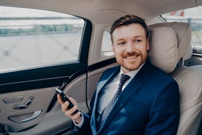 Smiling businessman holding mobile phone sitting at car