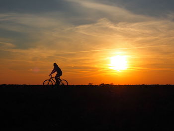 Silhouette person riding bicycle on field against sky during sunset
