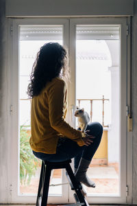 Full body side view of tranquil female in casual clothes sitting on stool near window and holding little kitten on knees while chilling at home