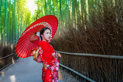 Mid adult woman holding red umbrella on footpath amidst bamboo forest