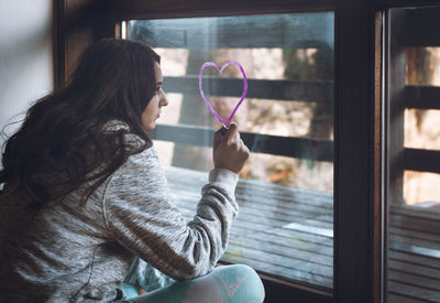 Woman drawing pink heart shape on glass window while sitting at home