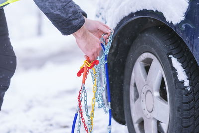 Cropped hands of man holding chain equipment by car tire during winter