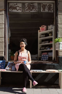 Happy owner having coffee while sitting on table by cushions outside fabric shop