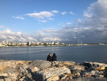 People on rocks by sea against sky in city