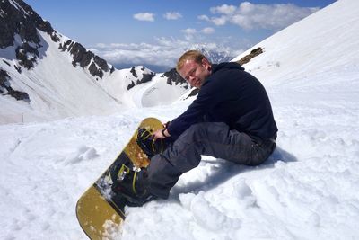 Side view of man sitting with snowboard on snowcapped mountain during sunny day