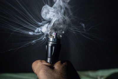 Cropped hand of person holding electronic cigarette with smoke against black background
