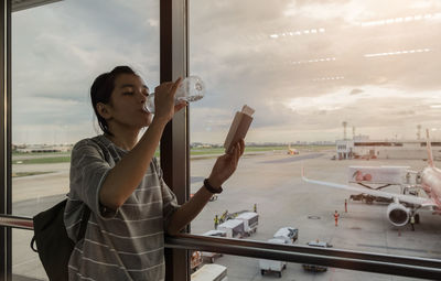 Woman with backpack drinking water from bottle while holding book standing against window at airport