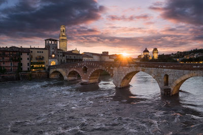 Bridge over river by buildings against cloudy sky during sunset