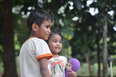 Thoughtful siblings smiling while standing against trees