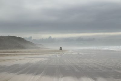 Mid distance view of car moving on beach against cloudy sky