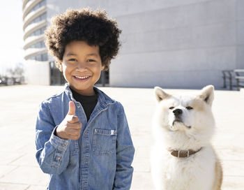 Smiling boy gesturing thumbs up while standing by dog sitting on footpath