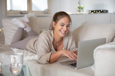 Smiling young woman using laptop while lying on sofa at home