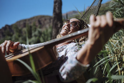 Woman with sunglasses laughing while playing guitar lying on a hammock