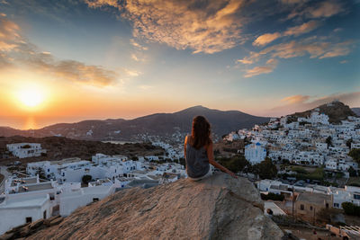 Rear view of woman looking at cityscape while sitting on rock against sky during sunset