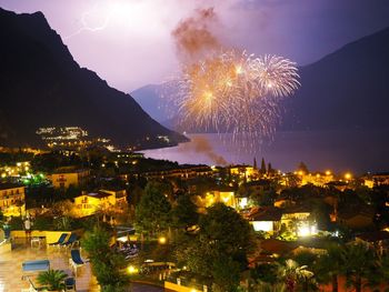 Fireworks in illuminated town by sea against sky at night