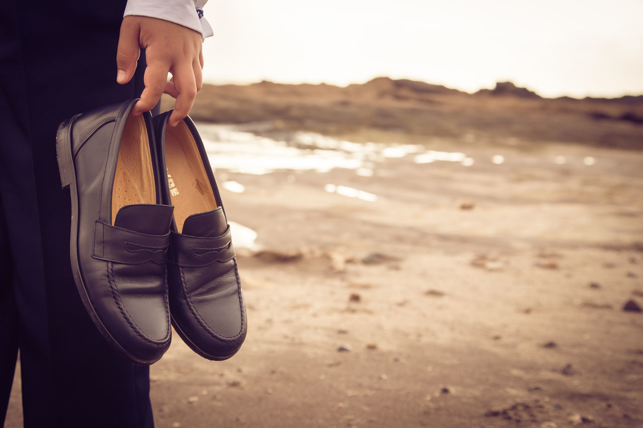 low section, beach, shoe, sand, person, lifestyles, standing, leisure activity, focus on foreground, men, footwear, close-up, shore, part of, sea, outdoors, day