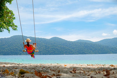 Woman swinging at beach against mountains