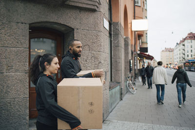 Male and female workers carrying cardboard boxes while walking in city during delivery