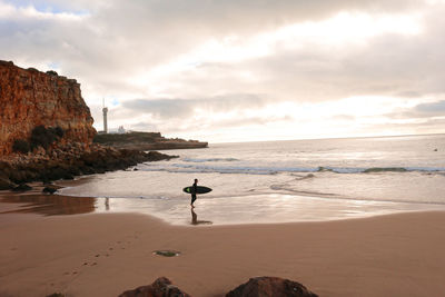 Full length of man holding surfboard walking on beach against sky