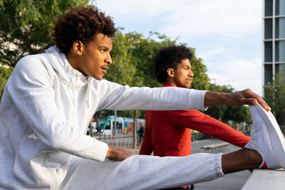 Side view of african american male doing hamstring stretch with leg on street fence near black friend