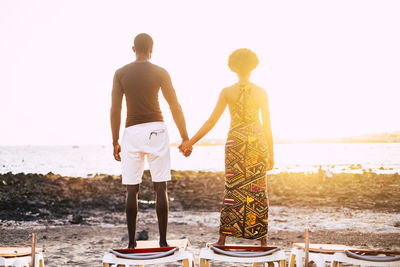 Rear view of couple holding hands while standing at beach against clear sky