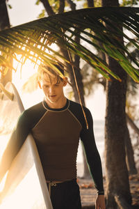 Young man carrying surfboard at beach during sunset