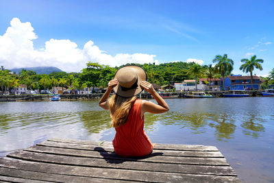 Beautiful young woman sitting on wooden pier enjoying relaxing sunny windy day in brazil.