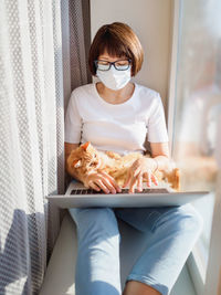 Woman in medical mask remote works from home with laptop and ginger cat. lockdown quarantine.