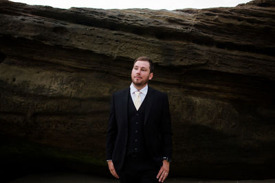 Groom posing in front of rock at beach in san diego