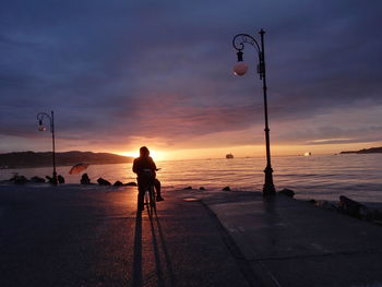 Silhouette woman on street by sea against sky during sunset