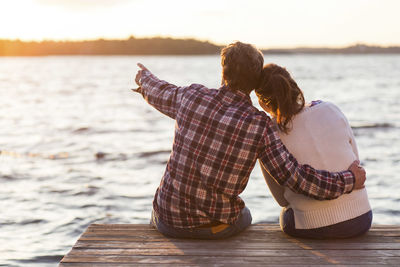 Rear view of man showing something to woman while sitting on pier against sea at sunset