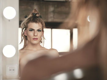 Shirtless transgender guy with makeup curling hair with rollers in front of illuminated mirror in studio