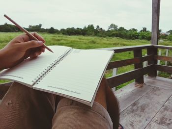 Low section of man writing on book in porch against grassy field