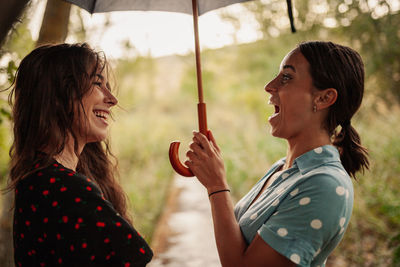 Side view of cheerful lesbian couple with umbrella standing outdoors