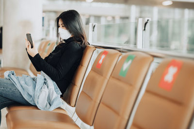 Woman wearing mask using smart phone sitting at airport